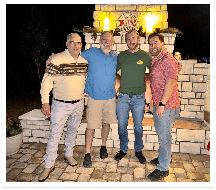 Four men smile for a photo in front of a cozy brick fireplace, creating a warm and inviting atmosphere.